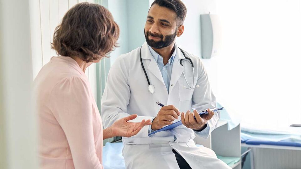 Male doctor with a stethoscope consulting an older woman in a clinic, holding a clipboard and pen as they talk.